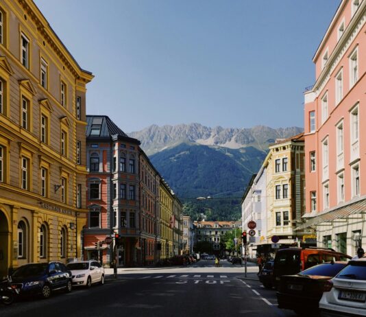 Autriche : un frein aux loyers pour tous-tes Rue dégagée en Autriche bordée de bâtiments anciens aux façades colorées – jaune, rose et ocre – avec des voitures stationnées le long du trottoir. Au fond, une chaîne de montagnes vertes coiffée de crêtes rocheuses se détache sous un ciel bleu clair. Sur la chaussée, le mot « STOP » est peint devant un carrefour signalé par un feu tricolore.