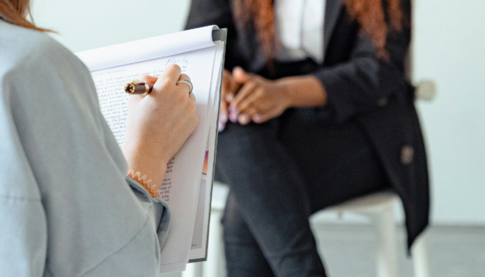 Personne de dos tenant un stylo et un bloc-notes, écrivant des notes manuscrites, en face d’une autre personne assise sur un tabouret, les mains jointes sur les genoux, vêtue d’un tailleur sombre.