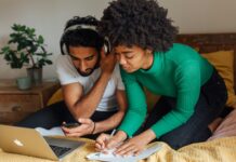 4 raisons de voter oui à l’imposition individuelle Image en gros plan d’un homme et d’une femme assis côte à côte sur un lit recouvert d’une couverture moutarde. L’homme, vêtu d’un t-shirt blanc et coiffé de cheveux noirs bouclés, porte un casque audio et tient un smartphone en main en regardant l’écran. La femme, penchée à ses côtés, porte un pull vert à côtes et a les cheveux crépus ; elle écrit au stylo sur une feuille de papier posée sur le lit. Devant eux, un ordinateur portable argenté est ouvert. À l’arrière-plan, une table de chevet en bois supporte une plante en pot et une tasse.