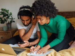 4 raisons de voter oui à l’imposition individuelle Image en gros plan d’un homme et d’une femme assis côte à côte sur un lit recouvert d’une couverture moutarde. L’homme, vêtu d’un t-shirt blanc et coiffé de cheveux noirs bouclés, porte un casque audio et tient un smartphone en main en regardant l’écran. La femme, penchée à ses côtés, porte un pull vert à côtes et a les cheveux crépus ; elle écrit au stylo sur une feuille de papier posée sur le lit. Devant eux, un ordinateur portable argenté est ouvert. À l’arrière-plan, une table de chevet en bois supporte une plante en pot et une tasse.