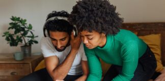 4 raisons de voter oui à l’imposition individuelle Image en gros plan d’un homme et d’une femme assis côte à côte sur un lit recouvert d’une couverture moutarde. L’homme, vêtu d’un t-shirt blanc et coiffé de cheveux noirs bouclés, porte un casque audio et tient un smartphone en main en regardant l’écran. La femme, penchée à ses côtés, porte un pull vert à côtes et a les cheveux crépus ; elle écrit au stylo sur une feuille de papier posée sur le lit. Devant eux, un ordinateur portable argenté est ouvert. À l’arrière-plan, une table de chevet en bois supporte une plante en pot et une tasse.