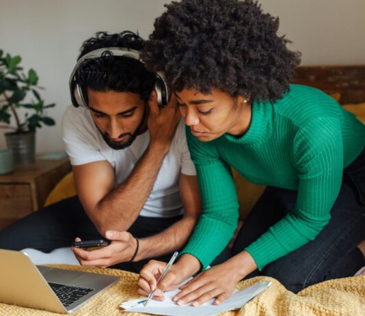 4 raisons de voter oui à l’imposition individuelle Image en gros plan d’un homme et d’une femme assis côte à côte sur un lit recouvert d’une couverture moutarde. L’homme, vêtu d’un t-shirt blanc et coiffé de cheveux noirs bouclés, porte un casque audio et tient un smartphone en main en regardant l’écran. La femme, penchée à ses côtés, porte un pull vert à côtes et a les cheveux crépus ; elle écrit au stylo sur une feuille de papier posée sur le lit. Devant eux, un ordinateur portable argenté est ouvert. À l’arrière-plan, une table de chevet en bois supporte une plante en pot et une tasse.