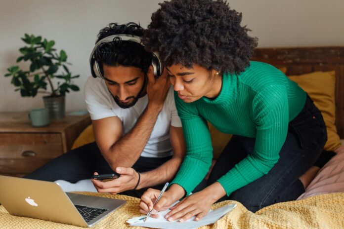 Image en gros plan d’un homme et d’une femme assis côte à côte sur un lit recouvert d’une couverture moutarde. L’homme, vêtu d’un t-shirt blanc et coiffé de cheveux noirs bouclés, porte un casque audio et tient un smartphone en main en regardant l’écran. La femme, penchée à ses côtés, porte un pull vert à côtes et a les cheveux crépus ; elle écrit au stylo sur une feuille de papier posée sur le lit. Devant eux, un ordinateur portable argenté est ouvert. À l’arrière-plan, une table de chevet en bois supporte une plante en pot et une tasse.