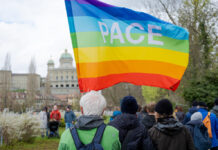 Marche de Pâques 2026 : Ensemble contre la guerre, la violence et l’oppression Au premier plan, une personne aux cheveux blancs, vêtue d'une veste verte et portant un sac à dos, tient un grand drapeau arc-en-ciel aux couleurs violette, bleue, verte, jaune, orange et rouge, sur lequel est inscrit le mot «PACE» (paix en italien). Autour d'elle, plusieurs participant·e·s habillés chaudement sont rassemblés dans un parc. En arrière-plan, on reconnaît les coupoles et la façade du Palais fédéral suisse, ainsi que des arbres aux feuilles naissantes, suggérant une journée de printemps nuageuse à Berne.