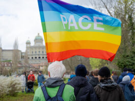 Marche de Pâques 2026 : Ensemble contre la guerre, la violence et l’oppression Au premier plan, une personne aux cheveux blancs, vêtue d'une veste verte et portant un sac à dos, tient un grand drapeau arc-en-ciel aux couleurs violette, bleue, verte, jaune, orange et rouge, sur lequel est inscrit le mot «PACE» (paix en italien). Autour d'elle, plusieurs participant·e·s habillés chaudement sont rassemblés dans un parc. En arrière-plan, on reconnaît les coupoles et la façade du Palais fédéral suisse, ainsi que des arbres aux feuilles naissantes, suggérant une journée de printemps nuageuse à Berne.