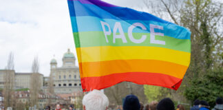 Marche de Pâques 2026 : Ensemble contre la guerre, la violence et l’oppression Au premier plan, une personne aux cheveux blancs, vêtue d'une veste verte et portant un sac à dos, tient un grand drapeau arc-en-ciel aux couleurs violette, bleue, verte, jaune, orange et rouge, sur lequel est inscrit le mot «PACE» (paix en italien). Autour d'elle, plusieurs participant·e·s habillés chaudement sont rassemblés dans un parc. En arrière-plan, on reconnaît les coupoles et la façade du Palais fédéral suisse, ainsi que des arbres aux feuilles naissantes, suggérant une journée de printemps nuageuse à Berne.
