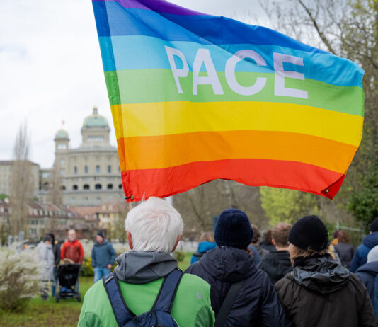 Marche de Pâques 2026 : Ensemble contre la guerre, la violence et l’oppression Au premier plan, une personne aux cheveux blancs, vêtue d'une veste verte et portant un sac à dos, tient un grand drapeau arc-en-ciel aux couleurs violette, bleue, verte, jaune, orange et rouge, sur lequel est inscrit le mot «PACE» (paix en italien). Autour d'elle, plusieurs participant·e·s habillés chaudement sont rassemblés dans un parc. En arrière-plan, on reconnaît les coupoles et la façade du Palais fédéral suisse, ainsi que des arbres aux feuilles naissantes, suggérant une journée de printemps nuageuse à Berne.
