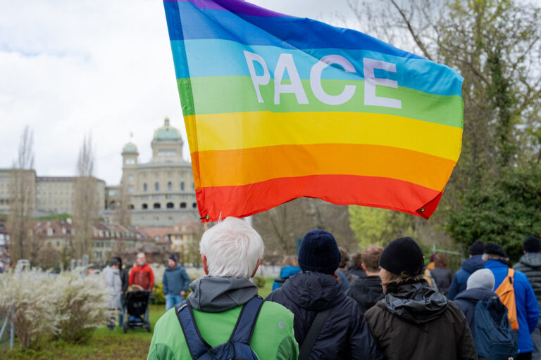 Au premier plan, une personne aux cheveux blancs, vêtue d'une veste verte et portant un sac à dos, tient un grand drapeau arc-en-ciel aux couleurs violette, bleue, verte, jaune, orange et rouge, sur lequel est inscrit le mot «PACE» (paix en italien). Autour d'elle, plusieurs participant·e·s habillés chaudement sont rassemblés dans un parc. En arrière-plan, on reconnaît les coupoles et la façade du Palais fédéral suisse, ainsi que des arbres aux feuilles naissantes, suggérant une journée de printemps nuageuse à Berne.