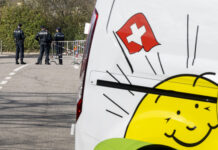 L’initiative de l’UDC plonge la Suisse dans le chaos Des policiers se tiennent près d’une barrière bloquant une route, tandis que le côté d’un véhicule blanc avec un drapeau suisse et un soleil souriant, le logo de l'UDC, est visible au premier plan.