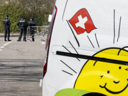 L’initiative de l’UDC plonge la Suisse dans le chaos Des policiers se tiennent près d’une barrière bloquant une route, tandis que le côté d’un véhicule blanc avec un drapeau suisse et un soleil souriant, le logo de l'UDC, est visible au premier plan.