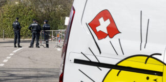 L’initiative de l’UDC plonge la Suisse dans le chaos Des policiers se tiennent près d’une barrière bloquant une route, tandis que le côté d’un véhicule blanc avec un drapeau suisse et un soleil souriant, le logo de l'UDC, est visible au premier plan.