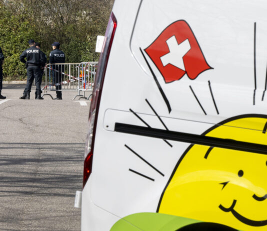 L’initiative de l’UDC plonge la Suisse dans le chaos Des policiers se tiennent près d’une barrière bloquant une route, tandis que le côté d’un véhicule blanc avec un drapeau suisse et un soleil souriant, le logo de l'UDC, est visible au premier plan.