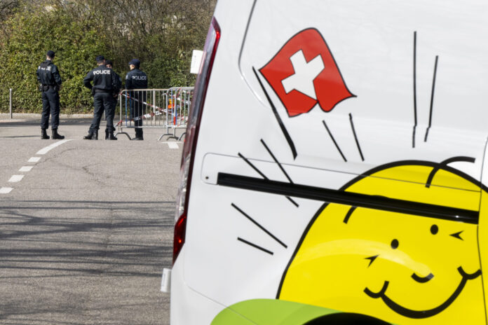 WAHLEN 2023, PARTEITAG, DV, DELEGIERTENVERSAMMLUNG, DELEGIERTE, SVP, Des policiers se tiennent près d’une barrière bloquant une route, tandis que le côté d’un véhicule blanc avec un drapeau suisse et un soleil souriant, le logo de l'UDC, est visible au premier plan.