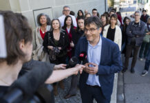 Deuxième tour au gouvernement vaudois : un choix évident Roger Nordmann, un homme portant des lunettes et une veste bleue est interviewé par une journaliste de la RTS (Radio Télévision Suisse) dans une rue. Il tient un micro tendu vers lui et s'exprime avec un léger sourire. En arrière-plan, une vingtaine de personnes se tiennent debout sur le trottoir, observant la scène.