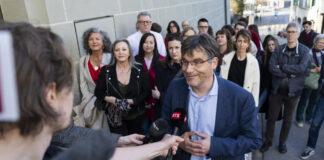 Deuxième tour au gouvernement vaudois : un choix évident Roger Nordmann, un homme portant des lunettes et une veste bleue est interviewé par une journaliste de la RTS (Radio Télévision Suisse) dans une rue. Il tient un micro tendu vers lui et s'exprime avec un léger sourire. En arrière-plan, une vingtaine de personnes se tiennent debout sur le trottoir, observant la scène.