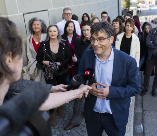 Deuxième tour au gouvernement vaudois : un choix évident Roger Nordmann, un homme portant des lunettes et une veste bleue est interviewé par une journaliste de la RTS (Radio Télévision Suisse) dans une rue. Il tient un micro tendu vers lui et s'exprime avec un léger sourire. En arrière-plan, une vingtaine de personnes se tiennent debout sur le trottoir, observant la scène.