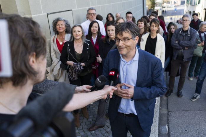 Roger Nordmann, un homme portant des lunettes et une veste bleue est interviewé par une journaliste de la RTS (Radio Télévision Suisse) dans une rue. Il tient un micro tendu vers lui et s'exprime avec un léger sourire. En arrière-plan, une vingtaine de personnes se tiennent debout sur le trottoir, observant la scène.