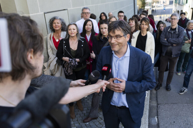 Roger Nordmann, un homme portant des lunettes et une veste bleue est interviewé par une journaliste de la RTS (Radio Télévision Suisse) dans une rue. Il tient un micro tendu vers lui et s'exprime avec un léger sourire. En arrière-plan, une vingtaine de personnes se tiennent debout sur le trottoir, observant la scène.