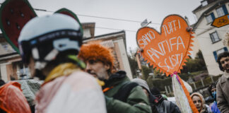Bonnes nouvelles en mars : « No Kings » et le carnaval antifasciste, Pedro Sánchez reste ferme, le service public médiatique reste intact et le PS progresse Une foule de manifestants réunis dans une rue urbaine par temps couvert. Au premier plan, une personne porte un casque décoré et une perruque orange. Au centre, un homme en veste verte tient une pancarte artisanale en forme de cœur orange avec des pompons, sur laquelle est écrit en italien « Siamo tutti antifa scisti » (« Nous sommes tous antifascistes »). Des rubans colorés sont attachés à la pancarte. En arrière-plan, on aperçoit des immeubles et d'autres participants au rassemblement.