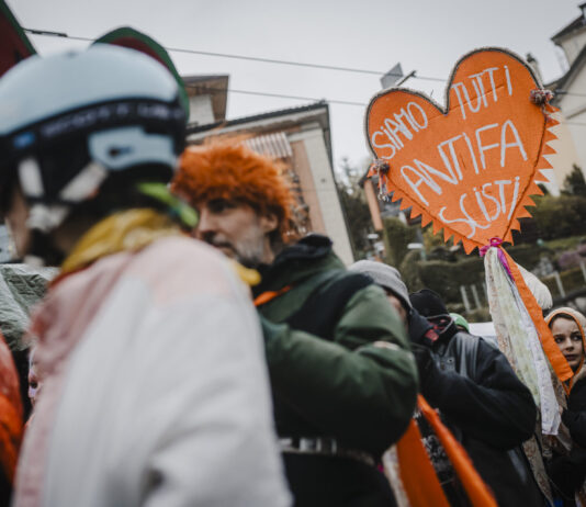 Bonnes nouvelles en mars : « No Kings » et le carnaval antifasciste, Pedro Sánchez reste ferme, le service public médiatique reste intact et le PS progresse Une foule de manifestants réunis dans une rue urbaine par temps couvert. Au premier plan, une personne porte un casque décoré et une perruque orange. Au centre, un homme en veste verte tient une pancarte artisanale en forme de cœur orange avec des pompons, sur laquelle est écrit en italien « Siamo tutti antifa scisti » (« Nous sommes tous antifascistes »). Des rubans colorés sont attachés à la pancarte. En arrière-plan, on aperçoit des immeubles et d'autres participants au rassemblement.