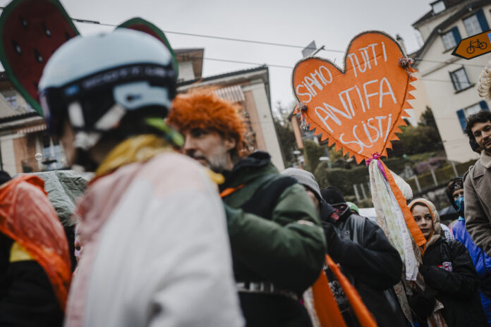 Une foule de manifestants réunis dans une rue urbaine par temps couvert. Au premier plan, une personne porte un casque décoré et une perruque orange. Au centre, un homme en veste verte tient une pancarte artisanale en forme de cœur orange avec des pompons, sur laquelle est écrit en italien « Siamo tutti antifa scisti » (« Nous sommes tous antifascistes »). Des rubans colorés sont attachés à la pancarte. En arrière-plan, on aperçoit des immeubles et d'autres participants au rassemblement.