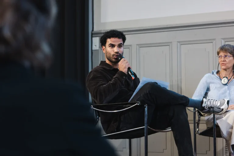 Un jeune homme aux cheveux bouclés, vêtu d'une veste noire, est assis sur une chaise et tient un microphone, semblant s'exprimer lors d'un événement public tel qu'une conférence ou un panel de discussion. Il tient des documents dans l'autre main. À sa droite, une femme aux cheveux gris, portant un haut bleu clair, est assise et l'écoute. En arrière-plan, on aperçoit un écran de projection et des panneaux gris. Au premier plan, une silhouette floue est visible, suggérant la présence d'un public.