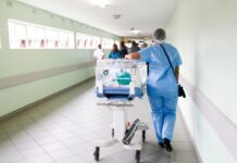 Initiative du chaos : les syndicats et la gauche lancent leur campagne Une professionnelle de santé vêtue de blouse et pantalon bleus, portant un filet de protection sur les cheveux, pousse un incubateur mobile transparent contenant un nourrisson le long d'un couloir hospitalier lumineux. D'autres personnes sont visibles en arrière-plan dans ce même couloir aux murs vert clair.