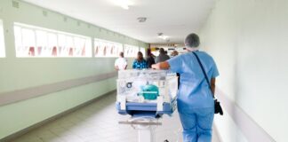 Initiative du chaos : les syndicats et la gauche lancent leur campagne Une professionnelle de santé vêtue de blouse et pantalon bleus, portant un filet de protection sur les cheveux, pousse un incubateur mobile transparent contenant un nourrisson le long d'un couloir hospitalier lumineux. D'autres personnes sont visibles en arrière-plan dans ce même couloir aux murs vert clair.