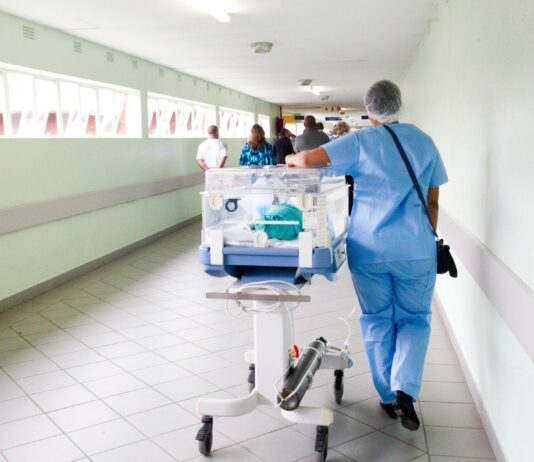 Initiative du chaos : les syndicats et la gauche lancent leur campagne Une professionnelle de santé vêtue de blouse et pantalon bleus, portant un filet de protection sur les cheveux, pousse un incubateur mobile transparent contenant un nourrisson le long d'un couloir hospitalier lumineux. D'autres personnes sont visibles en arrière-plan dans ce même couloir aux murs vert clair.