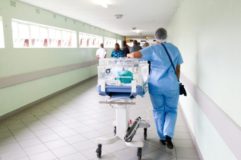 Une professionnelle de santé vêtue de blouse et pantalon bleus, portant un filet de protection sur les cheveux, pousse un incubateur mobile transparent contenant un nourrisson le long d'un couloir hospitalier lumineux. D'autres personnes sont visibles en arrière-plan dans ce même couloir aux murs vert clair.