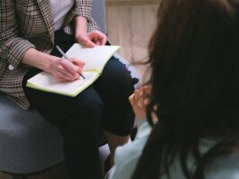 300 000 francs pour l’aide aux victimes : le Parlement corrige une décision désastreuse Une professionnelle portant une veste à carreaux prend des notes avec un stylo dans un carnet ouvert posé sur ses genoux, assise face à une personne aux cheveux bruns vue de dos, dans ce qui semble être un entretien ou une séance de consultation.