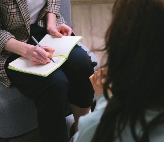300 000 francs pour l’aide aux victimes : le Parlement corrige une décision désastreuse Une professionnelle portant une veste à carreaux prend des notes avec un stylo dans un carnet ouvert posé sur ses genoux, assise face à une personne aux cheveux bruns vue de dos, dans ce qui semble être un entretien ou une séance de consultation.