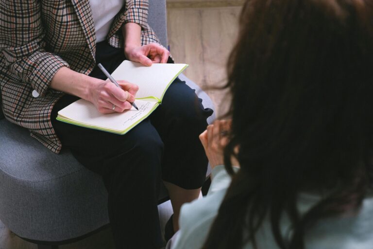 Une professionnelle portant une veste à carreaux prend des notes avec un stylo dans un carnet ouvert posé sur ses genoux, assise face à une personne aux cheveux bruns vue de dos, dans ce qui semble être un entretien ou une séance de consultation.