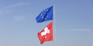 Accords bilatéraux en danger : ce que l’initiative du chaos de l’UDC changerait pour vous Deux drapeaux hissés sur un même mât par une journée ensoleillée. En haut, le drapeau de l'Union européenne — fond bleu orné d'un cercle de douze étoiles dorées. En dessous, le drapeau de la Suisse — fond rouge avec une croix blanche centrale. À l'arrière-plan, un lac calme, une ligne de collines verdoyantes et une ville aux maisons colorées, sous un ciel bleu légèrement nuageux.