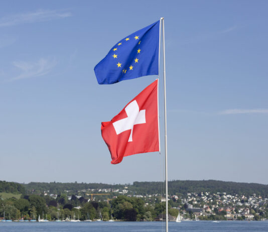 Accords bilatéraux en danger : ce que l’initiative du chaos de l’UDC changerait pour vous Deux drapeaux hissés sur un même mât par une journée ensoleillée. En haut, le drapeau de l'Union européenne — fond bleu orné d'un cercle de douze étoiles dorées. En dessous, le drapeau de la Suisse — fond rouge avec une croix blanche centrale. À l'arrière-plan, un lac calme, une ligne de collines verdoyantes et une ville aux maisons colorées, sous un ciel bleu légèrement nuageux.