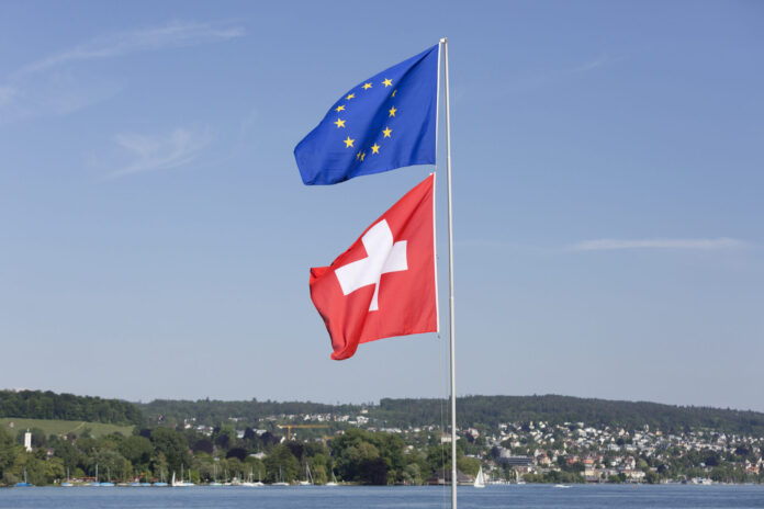 Deux drapeaux hissés sur un même mât par une journée ensoleillée. En haut, le drapeau de l'Union européenne — fond bleu orné d'un cercle de douze étoiles dorées. En dessous, le drapeau de la Suisse — fond rouge avec une croix blanche centrale. À l'arrière-plan, un lac calme, une ligne de collines verdoyantes et une ville aux maisons colorées, sous un ciel bleu légèrement nuageux.