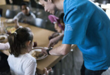 Le service civil : un acquis compliqué à obtenir, devenu essentiel Vue rapprochée d'un réfectoire où un volontaire du service civil, identifiable par son t-shirt bleu portant l'inscription "Service civil" en plusieurs langues (français, italien, allemand et romanche), sert de la nourriture à une jeune fille. La jeune fille, assise à une table, tend son assiette pour recevoir sa portion. Autour d'elle, d'autres personnes sont assises et partagent un repas. L'ambiance semble conviviale et chaleureuse, avec des plats variés visibles sur la table. L'image met en lumière l'engagement des volontaires du service civil dans des actions sociales et communautaires.