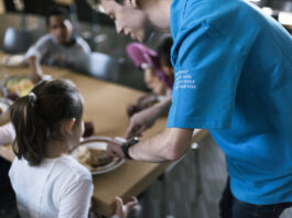Le service civil : un acquis compliqué à obtenir, devenu essentiel Vue rapprochée d'un réfectoire où un volontaire du service civil, identifiable par son t-shirt bleu portant l'inscription "Service civil" en plusieurs langues (français, italien, allemand et romanche), sert de la nourriture à une jeune fille. La jeune fille, assise à une table, tend son assiette pour recevoir sa portion. Autour d'elle, d'autres personnes sont assises et partagent un repas. L'ambiance semble conviviale et chaleureuse, avec des plats variés visibles sur la table. L'image met en lumière l'engagement des volontaires du service civil dans des actions sociales et communautaires.