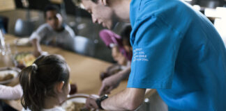 Le service civil : un acquis compliqué à obtenir, devenu essentiel Vue rapprochée d'un réfectoire où un volontaire du service civil, identifiable par son t-shirt bleu portant l'inscription "Service civil" en plusieurs langues (français, italien, allemand et romanche), sert de la nourriture à une jeune fille. La jeune fille, assise à une table, tend son assiette pour recevoir sa portion. Autour d'elle, d'autres personnes sont assises et partagent un repas. L'ambiance semble conviviale et chaleureuse, avec des plats variés visibles sur la table. L'image met en lumière l'engagement des volontaires du service civil dans des actions sociales et communautaires.