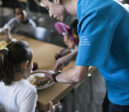 Le service civil : un acquis compliqué à obtenir, devenu essentiel Vue rapprochée d'un réfectoire où un volontaire du service civil, identifiable par son t-shirt bleu portant l'inscription "Service civil" en plusieurs langues (français, italien, allemand et romanche), sert de la nourriture à une jeune fille. La jeune fille, assise à une table, tend son assiette pour recevoir sa portion. Autour d'elle, d'autres personnes sont assises et partagent un repas. L'ambiance semble conviviale et chaleureuse, avec des plats variés visibles sur la table. L'image met en lumière l'engagement des volontaires du service civil dans des actions sociales et communautaires.