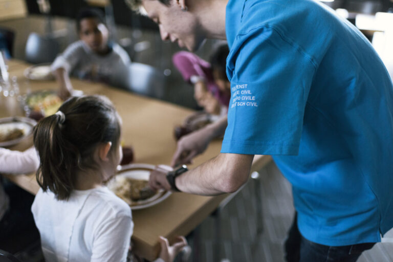 Vue rapprochée d'un réfectoire où un volontaire du service civil, identifiable par son t-shirt bleu portant l'inscription "Service civil" en plusieurs langues (français, italien, allemand et romanche), sert de la nourriture à une jeune fille. La jeune fille, assise à une table, tend son assiette pour recevoir sa portion. Autour d'elle, d'autres personnes sont assises et partagent un repas. L'ambiance semble conviviale et chaleureuse, avec des plats variés visibles sur la table. L'image met en lumière l'engagement des volontaires du service civil dans des actions sociales et communautaires.