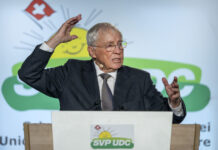 Une manœuvre stratégique : Blocher agrandit son empire médiatique Christoph Blocher, un homme âgé portant des lunettes se tient à un pupitre et gesticule vivement lors d'un discours. Il porte un costume sombre avec une cravate. Le pupitre est marqué du logo de l'UDC SVP, un parti politique suisse. En arrière-plan, on aperçoit le symbole des armoiries suisses (croix rouge sur fond blanc) et des éléments de design verts. L'homme semble parler avec engagement et souligne ses paroles par des gestes expressifs des mains. L'atmosphère paraît professionnelle et officielle, typique d'un événement politique ou d'un congrès de parti.