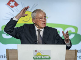 Une manœuvre stratégique : Blocher agrandit son empire médiatique Christoph Blocher, un homme âgé portant des lunettes se tient à un pupitre et gesticule vivement lors d'un discours. Il porte un costume sombre avec une cravate. Le pupitre est marqué du logo de l'UDC SVP, un parti politique suisse. En arrière-plan, on aperçoit le symbole des armoiries suisses (croix rouge sur fond blanc) et des éléments de design verts. L'homme semble parler avec engagement et souligne ses paroles par des gestes expressifs des mains. L'atmosphère paraît professionnelle et officielle, typique d'un événement politique ou d'un congrès de parti.