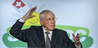 Une manœuvre stratégique : Blocher agrandit son empire médiatique Christoph Blocher, un homme âgé portant des lunettes se tient à un pupitre et gesticule vivement lors d'un discours. Il porte un costume sombre avec une cravate. Le pupitre est marqué du logo de l'UDC SVP, un parti politique suisse. En arrière-plan, on aperçoit le symbole des armoiries suisses (croix rouge sur fond blanc) et des éléments de design verts. L'homme semble parler avec engagement et souligne ses paroles par des gestes expressifs des mains. L'atmosphère paraît professionnelle et officielle, typique d'un événement politique ou d'un congrès de parti.