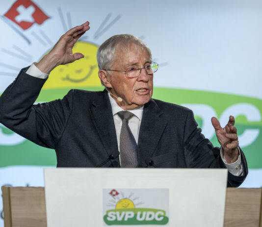 Une manœuvre stratégique : Blocher agrandit son empire médiatique Christoph Blocher, un homme âgé portant des lunettes se tient à un pupitre et gesticule vivement lors d'un discours. Il porte un costume sombre avec une cravate. Le pupitre est marqué du logo de l'UDC SVP, un parti politique suisse. En arrière-plan, on aperçoit le symbole des armoiries suisses (croix rouge sur fond blanc) et des éléments de design verts. L'homme semble parler avec engagement et souligne ses paroles par des gestes expressifs des mains. L'atmosphère paraît professionnelle et officielle, typique d'un événement politique ou d'un congrès de parti.