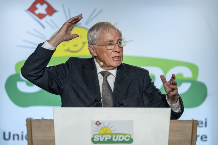 Christoph Blocher, un homme âgé portant des lunettes se tient à un pupitre et gesticule vivement lors d'un discours. Il porte un costume sombre avec une cravate. Le pupitre est marqué du logo de l'UDC SVP, un parti politique suisse. En arrière-plan, on aperçoit le symbole des armoiries suisses (croix rouge sur fond blanc) et des éléments de design verts. L'homme semble parler avec engagement et souligne ses paroles par des gestes expressifs des mains. L'atmosphère paraît professionnelle et officielle, typique d'un événement politique ou d'un congrès de parti.