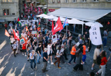 1er mai : les syndicats mettent en garde contre l’initiative du chaos Vue en plongée d'une manifestation de rue dans ce qui semble être une ville suisse ou française. Des centaines de personnes marchent sur des pavés, brandissant des drapeaux rouges, des drapeaux palestiniens et un drapeau aux couleurs transgenres (rose, blanc et bleu). Des pancartes sont visibles, dont l'une indique "No Justice No Peace" et une autre "Droit à l'éducation". Des terrasses de restaurants bordent la rue.