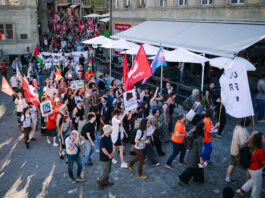 1er mai : les syndicats mettent en garde contre l’initiative du chaos Vue en plongée d'une manifestation de rue dans ce qui semble être une ville suisse ou française. Des centaines de personnes marchent sur des pavés, brandissant des drapeaux rouges, des drapeaux palestiniens et un drapeau aux couleurs transgenres (rose, blanc et bleu). Des pancartes sont visibles, dont l'une indique "No Justice No Peace" et une autre "Droit à l'éducation". Des terrasses de restaurants bordent la rue.