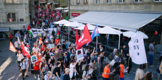 1er mai : les syndicats mettent en garde contre l’initiative du chaos Vue en plongée d'une manifestation de rue dans ce qui semble être une ville suisse ou française. Des centaines de personnes marchent sur des pavés, brandissant des drapeaux rouges, des drapeaux palestiniens et un drapeau aux couleurs transgenres (rose, blanc et bleu). Des pancartes sont visibles, dont l'une indique "No Justice No Peace" et une autre "Droit à l'éducation". Des terrasses de restaurants bordent la rue.