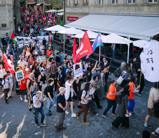 1er mai : les syndicats mettent en garde contre l’initiative du chaos Vue en plongée d'une manifestation de rue dans ce qui semble être une ville suisse ou française. Des centaines de personnes marchent sur des pavés, brandissant des drapeaux rouges, des drapeaux palestiniens et un drapeau aux couleurs transgenres (rose, blanc et bleu). Des pancartes sont visibles, dont l'une indique "No Justice No Peace" et une autre "Droit à l'éducation". Des terrasses de restaurants bordent la rue.